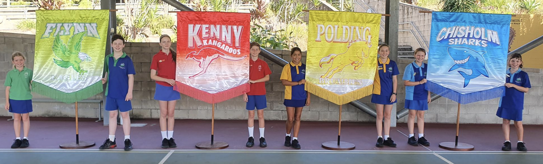 Students in school uniform standing in a hall holding colourful school house banners.