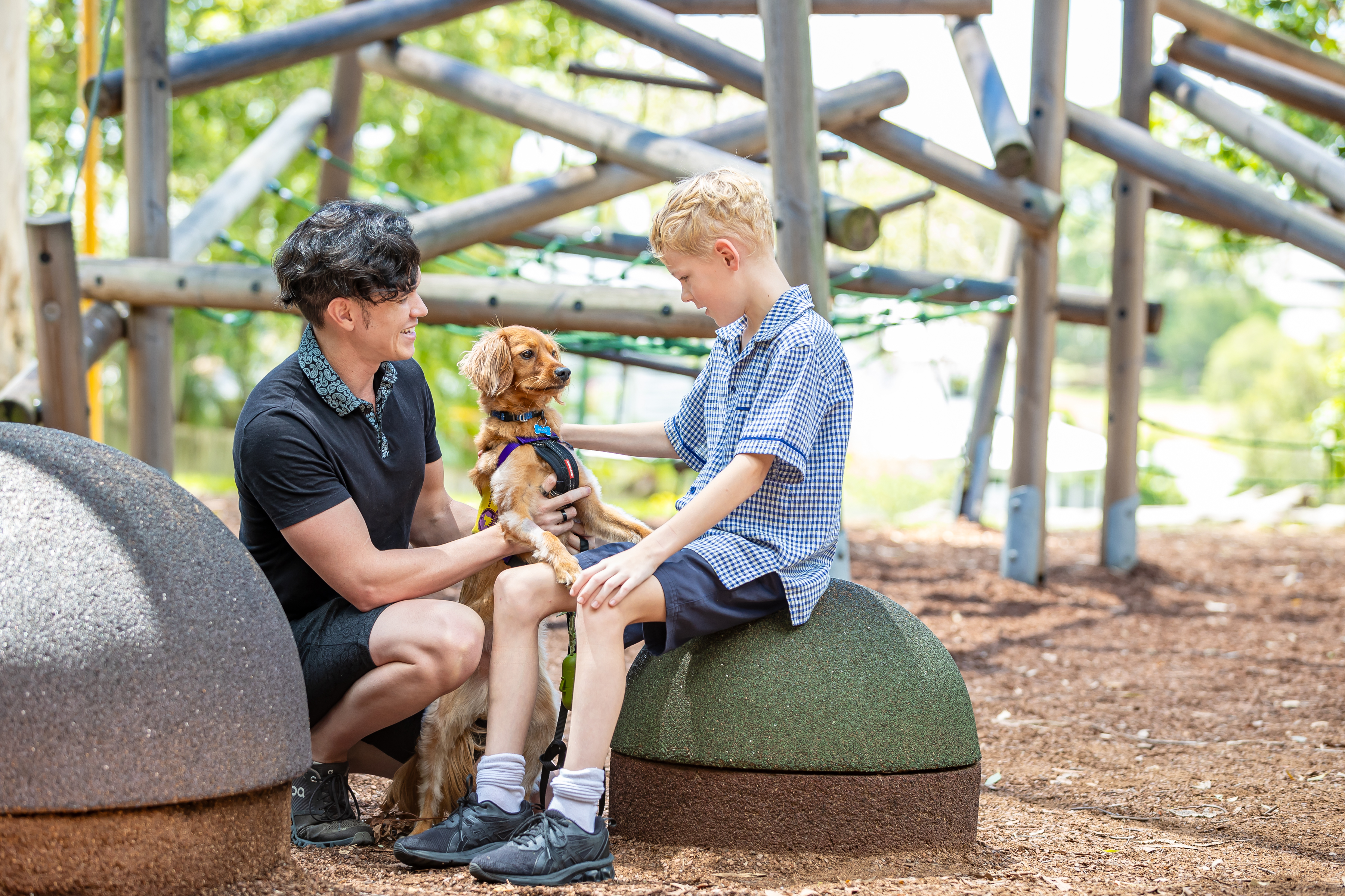 Student sitting outdoors with an adult and a small dog on a school playground.