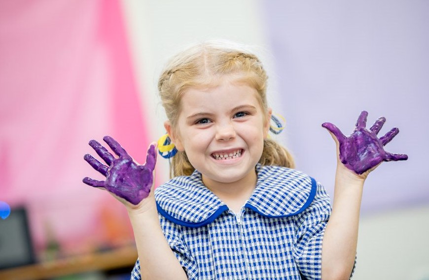 Young student in school uniform holding up hands covered in purple paint during an art activity.
