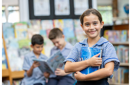 Student holding books in a classroom library, with other students reading in the background.